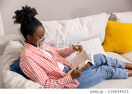 Smiling woman reading book while sitting on sofa at home. 110634425
