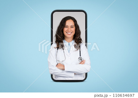 Cheerful young woman doctor in white uniform posing with stethoscope and folded arms in giant cellphone screen Cheerful young woman doctor in white uniform posing with stethoscope and folded arms in giant cellphone screen 110634697