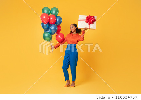 happy black teen girl holding balloons and present, yellow background 110634737