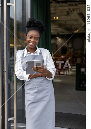 Dark-skinned young woman with notepad in hands standing in cafe door Dark-skinned young woman with notepad in hands standing in cafe door 110634835
