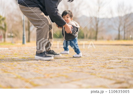 外でパパと一緒にあんよ（歩く）練習をして楽しそうな笑顔を見せる1歳の赤ちゃん 110635005