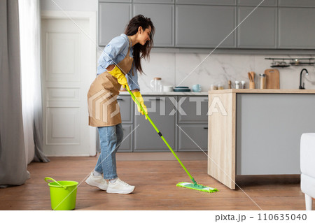 Focused woman in apron mopping floor with green bucket nearby Focused woman in apron mopping floor with green bucket nearby 110635040