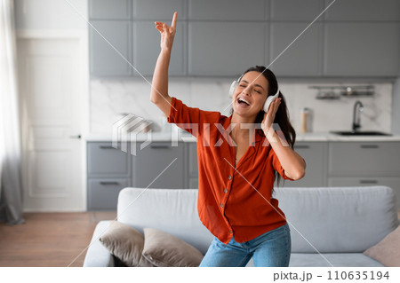 Joyful woman dancing and singing with headphones in a stylish living room Joyful woman dancing and singing with headphones in a stylish living room 110635194