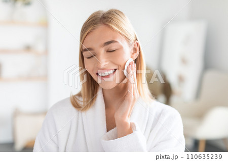 Smiling millennial woman moisturizing her face using cotton pad indoor 110635239