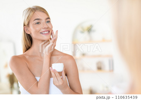 Pretty blonde lady applying moisturizer and enjoying moisturizing skincare indoors Pretty blonde lady applying moisturizer and enjoying moisturizing skincare indoors 110635439