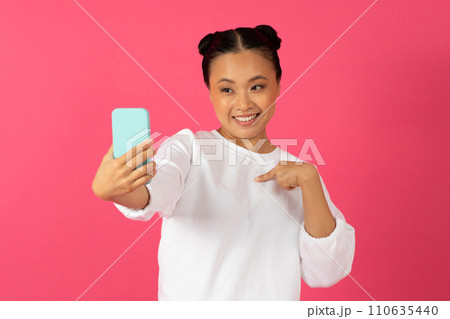 Happy young asian woman taking selfie on smartphone and pointing at herself 110635440