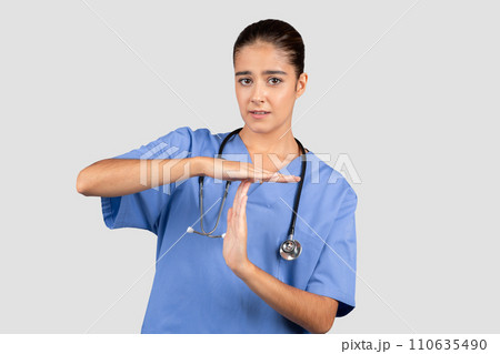 Concerned european nurse in blue scrubs making a timeout sign with her hands, indicating a need for a pause Concerned european nurse in blue scrubs making a timeout sign with her hands, indicating a need for a pause 110635490
