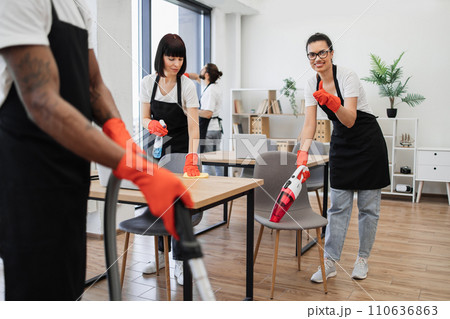 Multiethnic female cleaner vacuuming chair with portable vacuum cleaner. 110636863