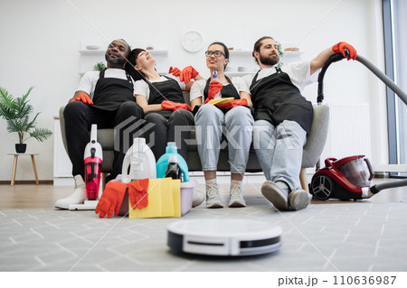 Team of housekeepers sitting on couch while robot vacuum cleaner cleans floor. 110636987