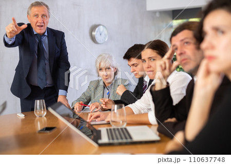 Outraged old manager getting mad at young adult mature office workers sitting with reports and laptops at table in meeting room Outraged old manager getting mad at young adult mature office workers sitting with reports and laptops at table in meeting room 110637748