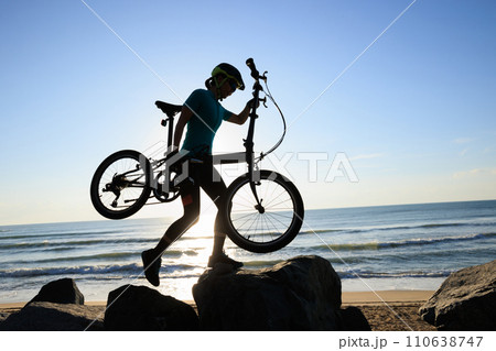 Woman carrying a folding bike on sunrise seaside road Woman carrying a folding bike on sunrise seaside road 110638747