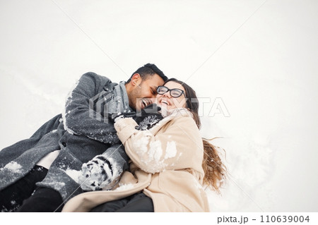 Indian man and caucasian woman walking in winter forest together. Lovely couple lay in snow. Man wearing grey coat and woman beige coat. Indian man and caucasian woman walking in winter forest together. Lovely couple lay in snow. Man wearing grey coat and woman beige coat. 110639004