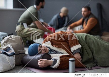 Side view portrait of Middle Eastern woman wearing hijab sleeping on floor at refugee shelter with people in background, copy space 110640368