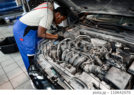 Young African auto mechanic checking car engine under the hood in auto service Young African auto mechanic checking car engine under the hood in auto service 110642070