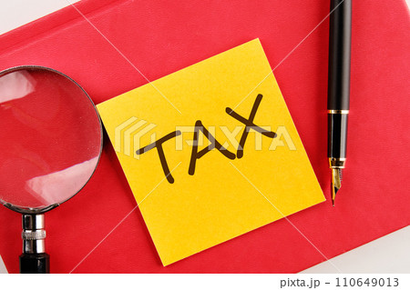 TAX the inscription on the sticker lying on a red notebook next to a magnifying glass and a fountain pen TAX the inscription on the sticker lying on a red notebook next to a magnifying glass and a fountain pen 110649013