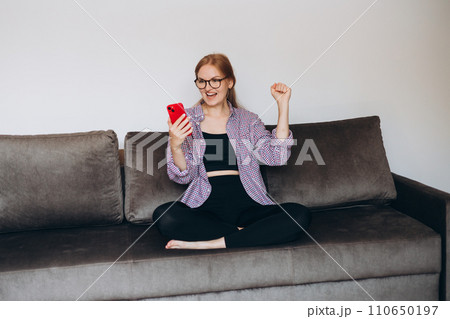 Satisfied young woman holding phone and celebrating on sofa at home. Smiling female sitting cross-legged on comfortable couch, happy lady relaxing with mobile phone in cozy living room, full body Satisfied young woman holding phone and celebrating on sofa at home. Smiling female sitting cross-legged on comfortable couch, happy lady relaxing with mobile phone in cozy living room, full body 110650197