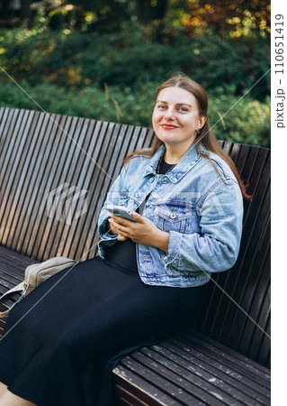 Positive cheerful woman tourist dressed in casual look holding smartphone gadget in hand for communicate on the city street. Happy woman chatting on cell smartphone and relaxing on the bench outdoors 110651419