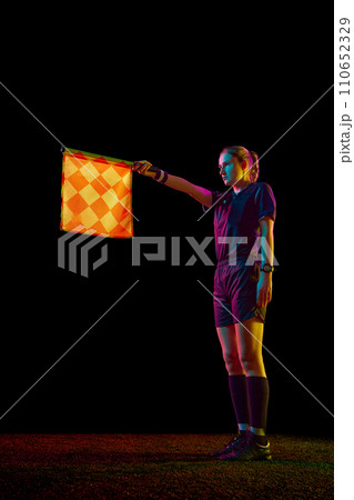 Young woman, assistant referee raising flag up meaning offside position against black studio background in neon light Young woman, assistant referee raising flag up meaning offside position against black studio background in neon light 110652329