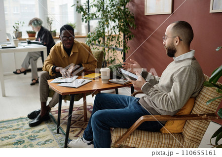 Biracial male coworkers discussing documents with infographics sitting at table in office Biracial male coworkers discussing documents with infographics sitting at table in office 110655161