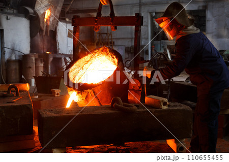 Industrial worker in protective gear pours molten metal at steel foundry. Manufacturing process in heavy industry with furnace. Skilled laborer handles high-temperature metallurgy, safety action. 110655455