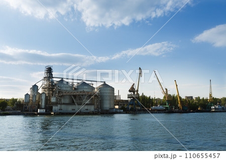 Industrial waterfront with grain elevator and cranes. Infrastructure for logistics, ship loading. Harbor dock panorama with heavy machinery against blue sky. Industrial waterfront with grain elevator and cranes. Infrastructure for logistics, ship loading. Harbor dock panorama with heavy machinery against blue sky. 110655457