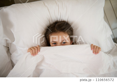 Top view of an adorable child girl lying on the bed, hiding half of her face under white blanket, looking at the camera 110656572