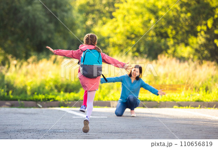 Back view of school girl running to mother after class on sunny street Back view of school girl running to mother after class on sunny street 110656819