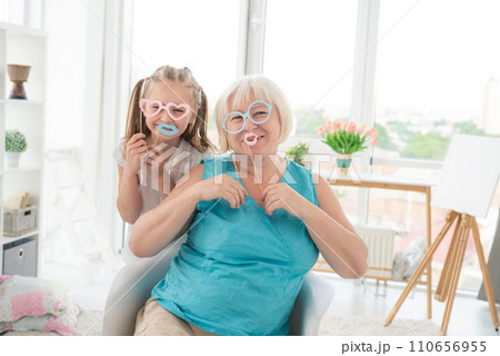 Smiling grandma having fun with cute granddaughter posing with paper glasses and lips at home 110656955