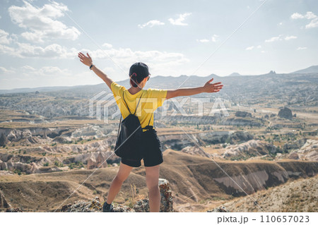 Woman tourist with backpack standing backwards on mountain top in Cappadocia with open arms, Turkey Woman tourist with backpack standing backwards on mountain top in Cappadocia with open arms, Turkey 110657023