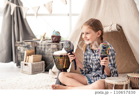 Smiling little girl with maracas in hands and ethnic drums on floor in children's room Smiling little girl with maracas in hands and ethnic drums on floor in children's room 110657830