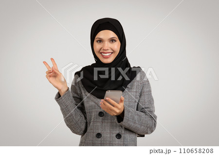 Happy Muslim woman with phone making peace sign 110658208
