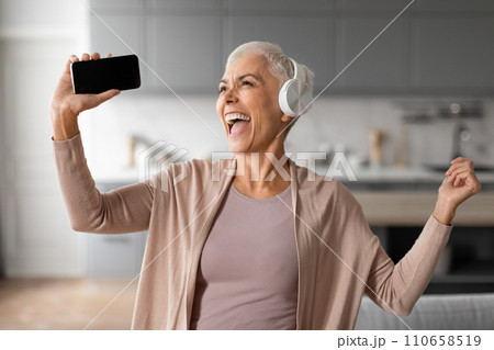senior woman with headset and phone dancing and singing indoor 110658519