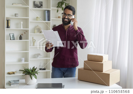 Indian businessman holding papers and speaking on cellphone, discussing order with customer 110658568