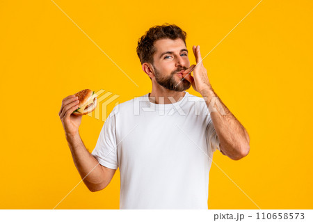 Portrait Of Smiling Hungry Man Eating Burger Licking Fingers, Studio Portrait Of Smiling Hungry Man Eating Burger Licking Fingers, Studio 110658573