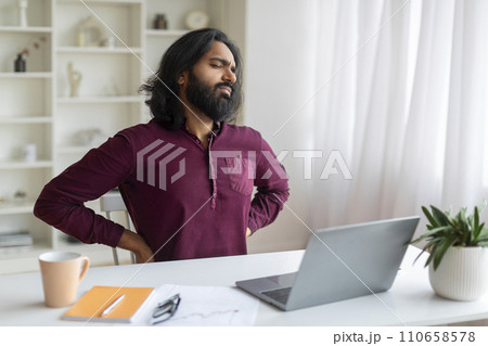 Indian Man Suffering From Backache While Sitting At Desk In Home Office Indian Man Suffering From Backache While Sitting At Desk In Home Office 110658578