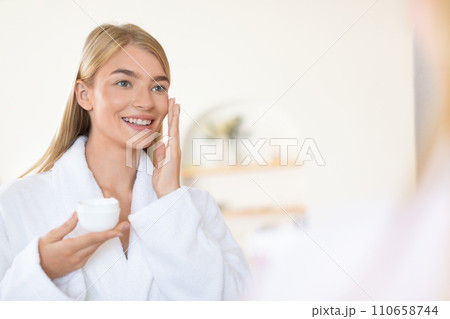 Attractive blonde young lady applies moisturizer in modern bathroom Attractive blonde young lady applies moisturizer in modern bathroom 110658744