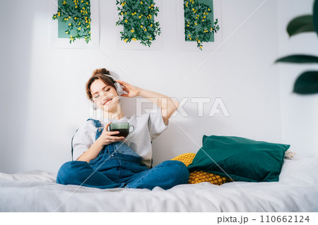 Woman with closed eyes listen music and enjoy cup of coffee or tea. Calm female spend free time at home enjoy favorite song with wireless modern headphones. Pastime weekend relax, no stress concept. 110662124