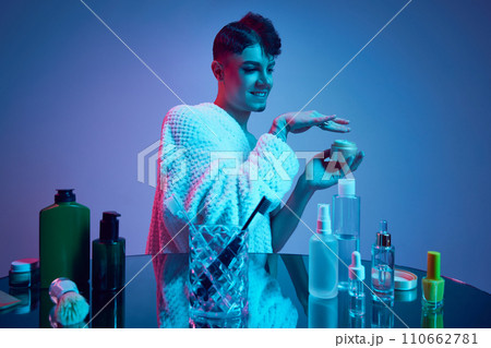 Young man with male and female appearance immerse in essence of self-care and cosmetic brilliance, while using cream. Young man with male and female appearance immerse in essence of self-care and cosmetic brilliance, while using cream. 110662781