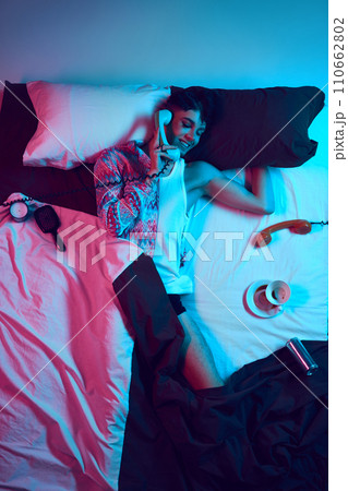 Gender fusion symphony. Visual overture. Creative portrait of young male model lying in bad and talking on retro phone as woman in neon light. 110662802