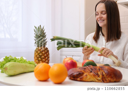 Cheerful joyful Caucasian brown haired woman wearing white shirt sitting at table in kitchen with fruit and vegetables holding leek smiling enjoying healthy nutrition Cheerful joyful Caucasian brown haired woman wearing white shirt sitting at table in kitchen with fruit and vegetables holding leek smiling enjoying healthy nutrition 110662855