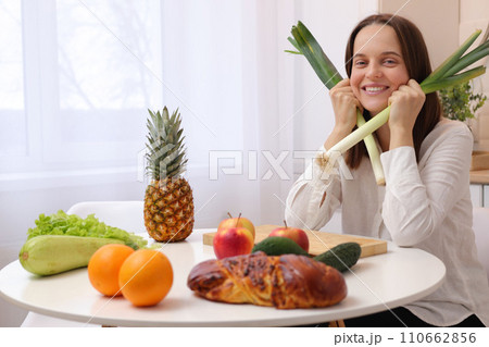 Pretty satisfied Caucasian brown haired woman wearing white shirt sitting at table in kitchen with fruit and vegetables preparing tasty healthy diet meal 110662856