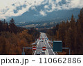 A Highway Through Fall Foliage with Mountains and Cloudy Skies in the Background 110662886