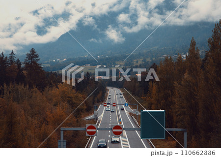 A Highway Through Fall Foliage with Mountains and Cloudy Skies in the Background 110662886