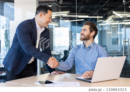Two professional businessmen in a modern office setting engaging in a friendly handshake, symbolizing a successful agreement or partnership. 110663323
