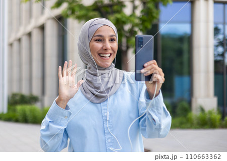 Smiling Arab businesswoman in hijab waving during a video call. Young female student or office worker outside modern building. 110663362