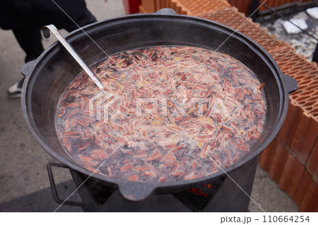 Round grill in the shape of a bowl with fire inside. A man prepares a hearth for barbecue. The round hearth is cleaned before cooking. The firewood lies in the bowl of the outdoor hearth. 110664254