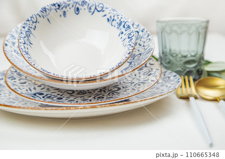 A set of plates of different sizes with a white middle and blue patterns on the side of the plate, stand on top of each other with a tilt, a fork and a spoon of gold - white color are located on the A set of plates of different sizes with a white middle and blue patterns on the side of the plate, stand on top of each other with a tilt, a fork and a spoon of gold - white color are located on the 110665348