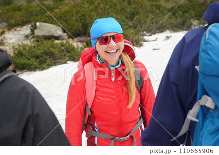 Young woman hiker in red jacket and sunglasses smiling to two other hikers, snow and trees behind her Young woman hiker in red jacket and sunglasses smiling to two other hikers, snow and trees behind her 110665906