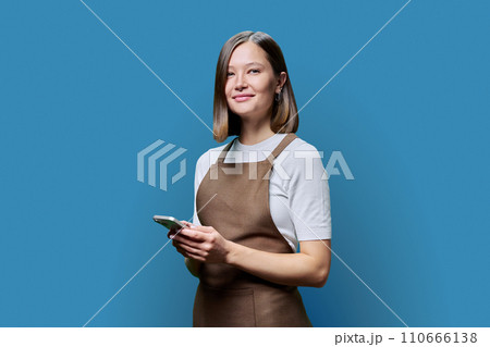 Portrait of young woman in apron holding smartphone in hands on blue background Portrait of young woman in apron holding smartphone in hands on blue background 110666138