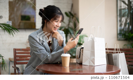 Young beautiful Asian female is using her phone in a cafe after her shopping day in the city. 110671504